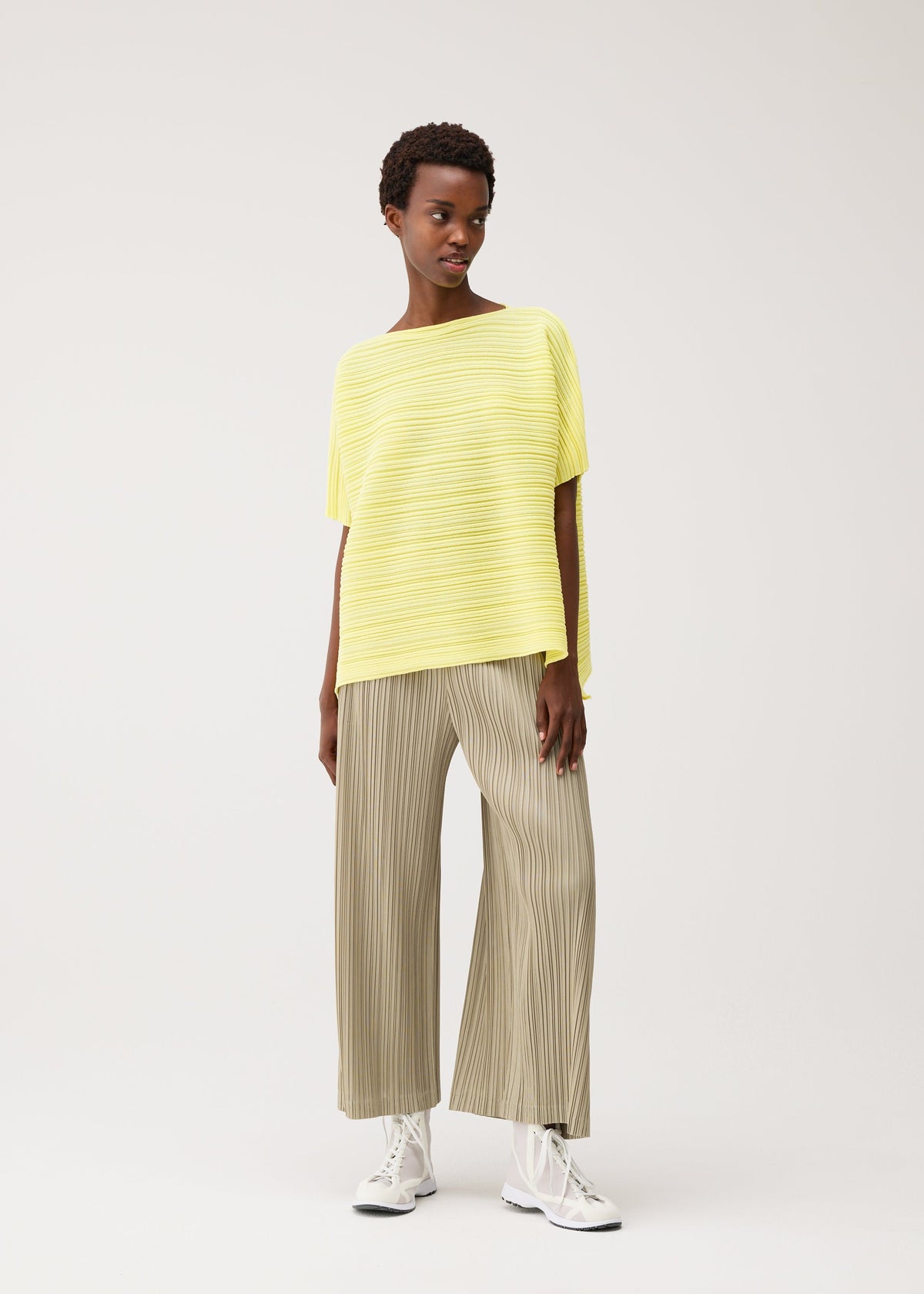 A woman poses against a plain background wearing the PAPER BOUNCE KNIT wide-leg beige pleated pants from PLEATS PLEASE ISSEY MIYAKE, paired with a loose yellow striped boat-neck top and white sneakers, looking to the side in a relaxed stance. view 8 of 8
