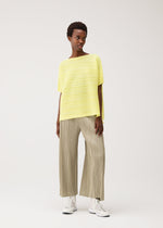 A woman poses against a plain background wearing the PAPER BOUNCE KNIT wide-leg beige pleated pants from PLEATS PLEASE ISSEY MIYAKE, paired with a loose yellow striped boat-neck top and white sneakers, looking to the side in a relaxed stance.