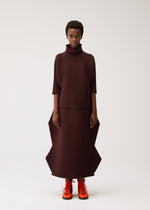 A person wearing the ZIG ZAG KNIT top and skirt from PLEATS PLEASE ISSEY MIYAKE, featuring sculptural shapes, paired with bright orange boots, stands facing forward against a plain white background.