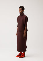 Against a plain light background, a person wears the PLEATS PLEASE ISSEY MIYAKE ZIG ZAG KNIT high-neck top and matching long skirt, paired with bright orange high-top sneakers.