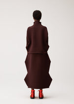 A person stands facing away, wearing the PLEATS PLEASE ISSEY MIYAKE ZIG ZAG KNIT—a structured, ribbed, dark brown seamless knit top featuring geometric patterns—with bright red shoes, set against a plain light background.