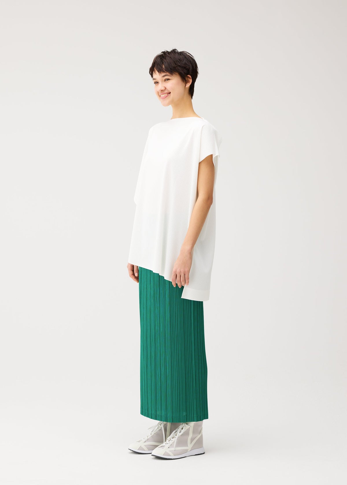 A person smiles against a plain background, wearing the loose white round-neck A-POC SLICE tunic and a long green pleated skirt by PLEATS PLEASE ISSEY MIYAKE, paired with white shoes. view 7 of 9