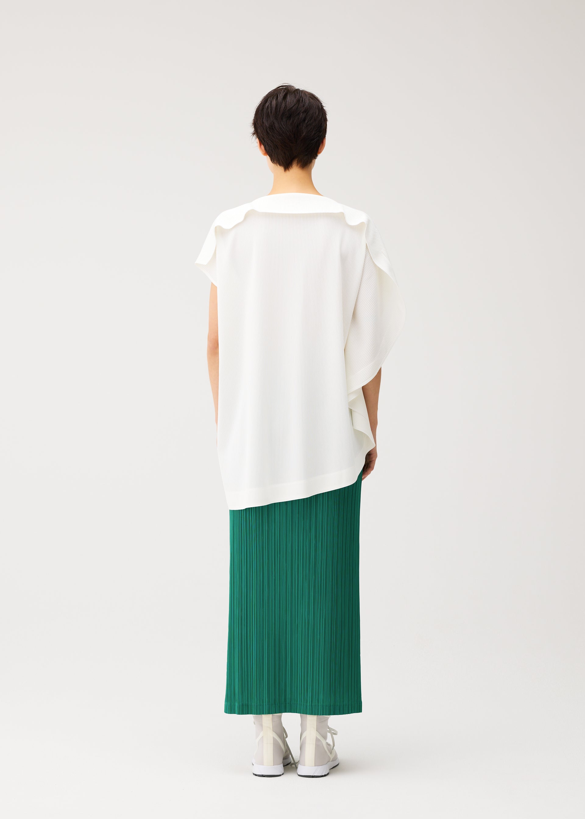 Zoomed view A person with short dark hair faces away, wearing the PLEATS PLEASE ISSEY MIYAKE A-POC SLICE loose round-neck tunic with an asymmetrical hem, a long green pleated skirt, and white shoes against a plain light background.