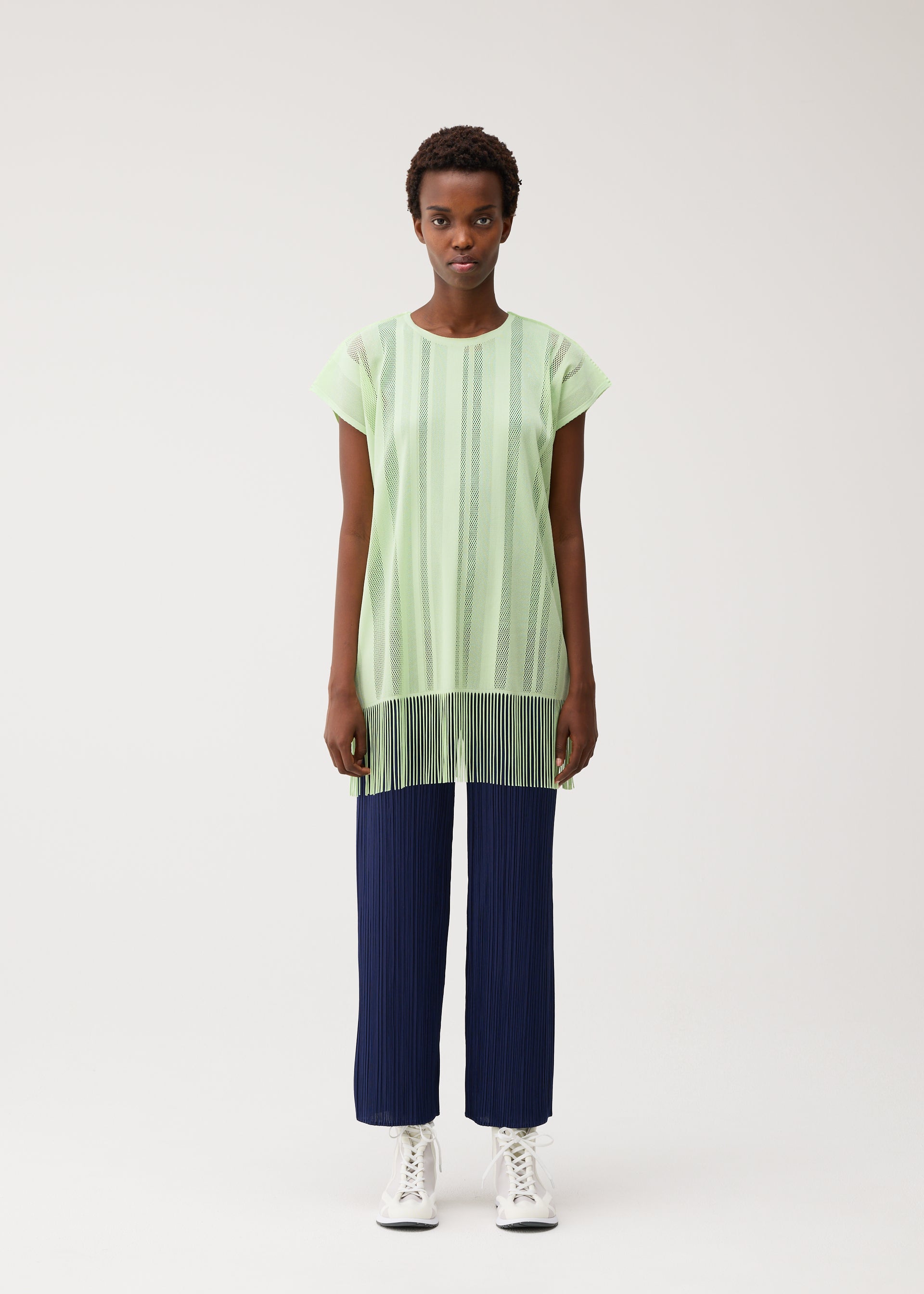 Zoomed view A person stands against a plain background, wearing the PLEATS PLEASE ISSEY MIYAKE A-POC LACE light green round-neck tunic with sheer vertical stripes and fringe made from mesh material, paired with dark blue pleated pants and white high-top sneakers.