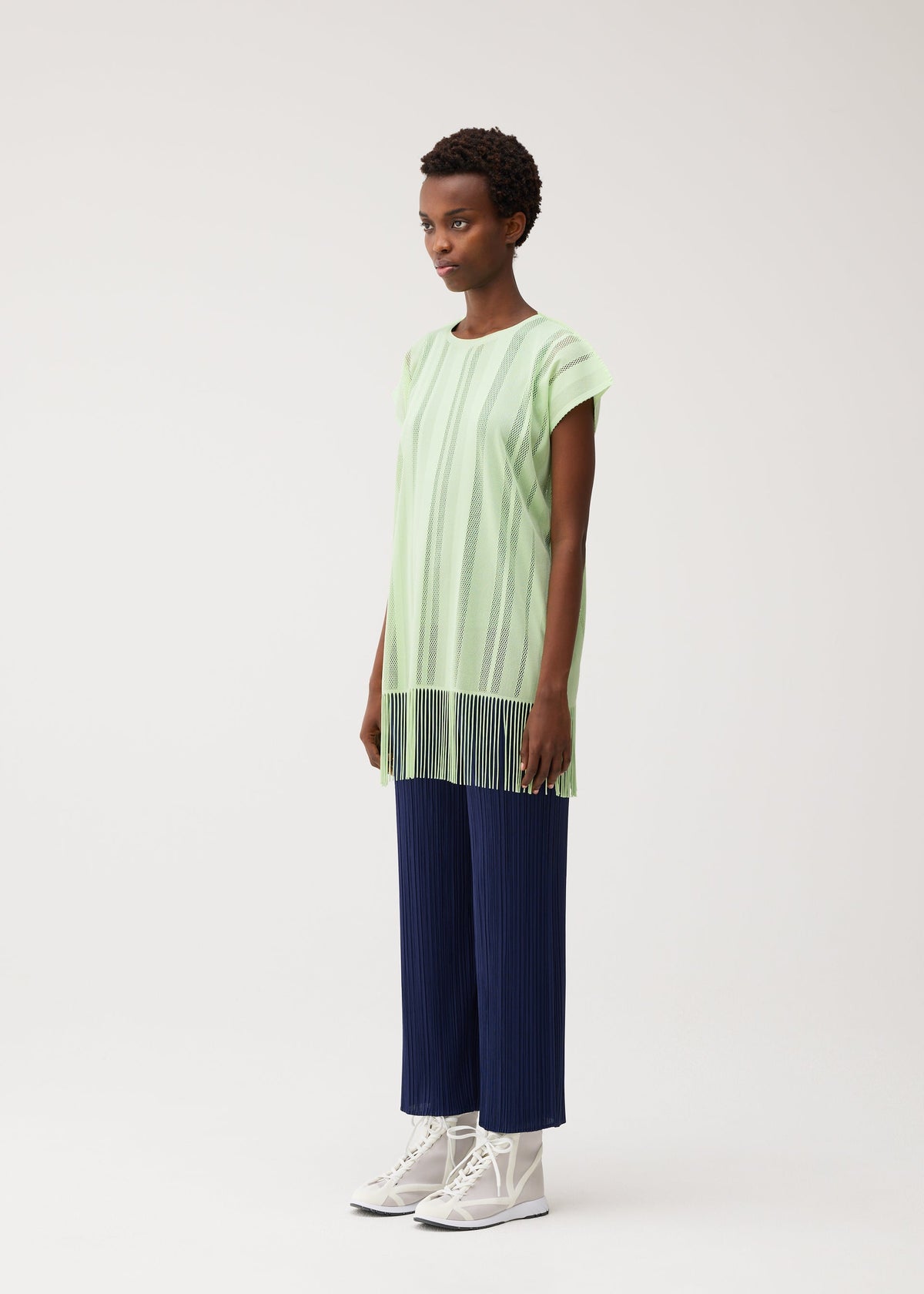 A person stands against a light background wearing the A-POC LACE tunic by PLEATS PLEASE ISSEY MIYAKE—a loose, light green fringed top with vertical stripes and fringe at the hem—paired with navy pleated wide-leg pants and white lace-up boots. view 6 of 9
