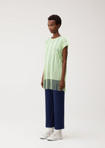 A person stands against a light background wearing the A-POC LACE tunic by PLEATS PLEASE ISSEY MIYAKE—a loose, light green fringed top with vertical stripes and fringe at the hem—paired with navy pleated wide-leg pants and white lace-up boots.