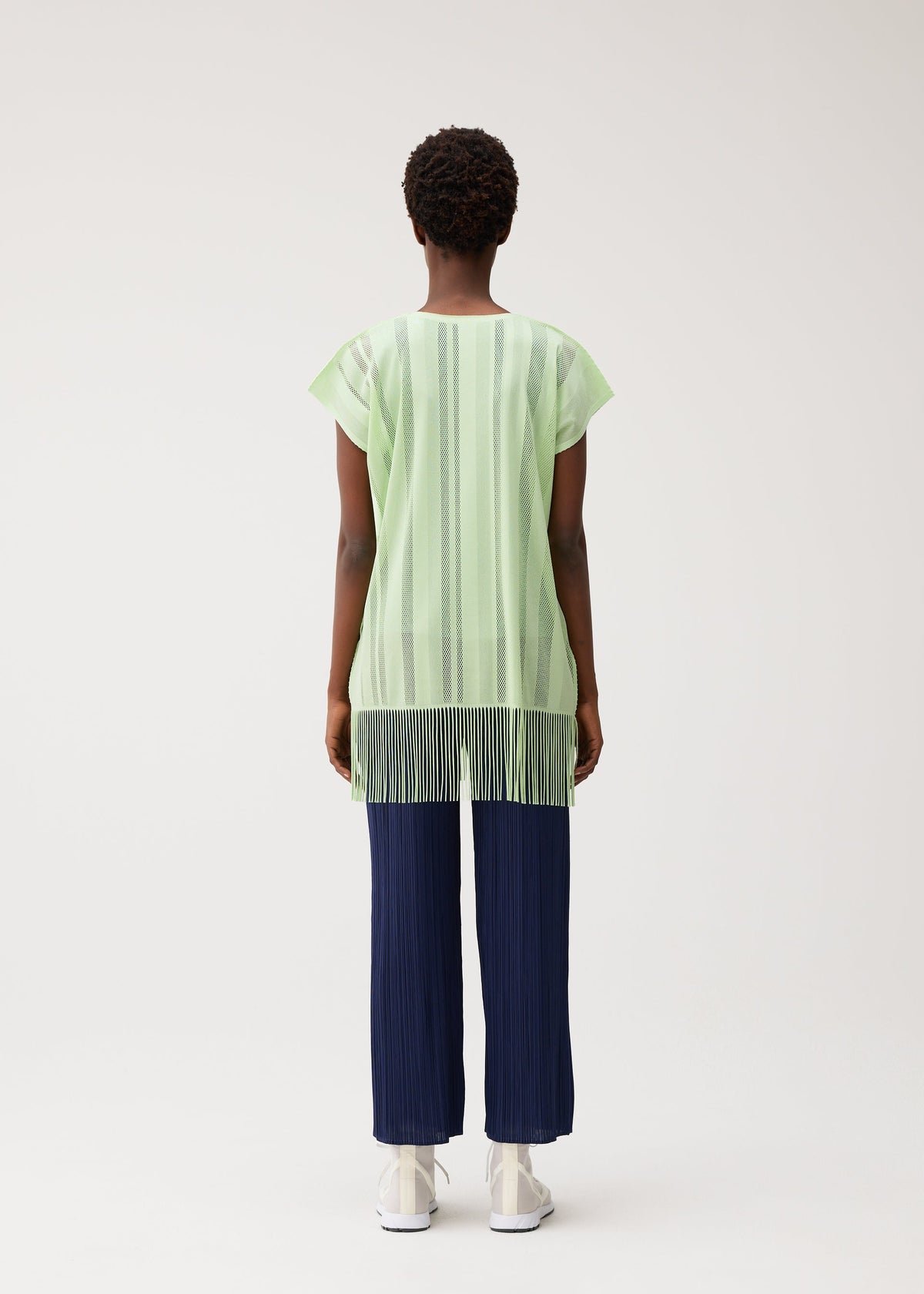 A person stands facing away against a plain light background, wearing the PLEATS PLEASE ISSEY MIYAKE A-POC LACE short-sleeve fringed tunic with vertical stripes in light green, paired with navy blue pleated pants and white shoes. view 7 of 9