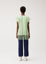 A person stands facing away against a plain light background, wearing the PLEATS PLEASE ISSEY MIYAKE A-POC LACE short-sleeve fringed tunic with vertical stripes in light green, paired with navy blue pleated pants and white shoes.