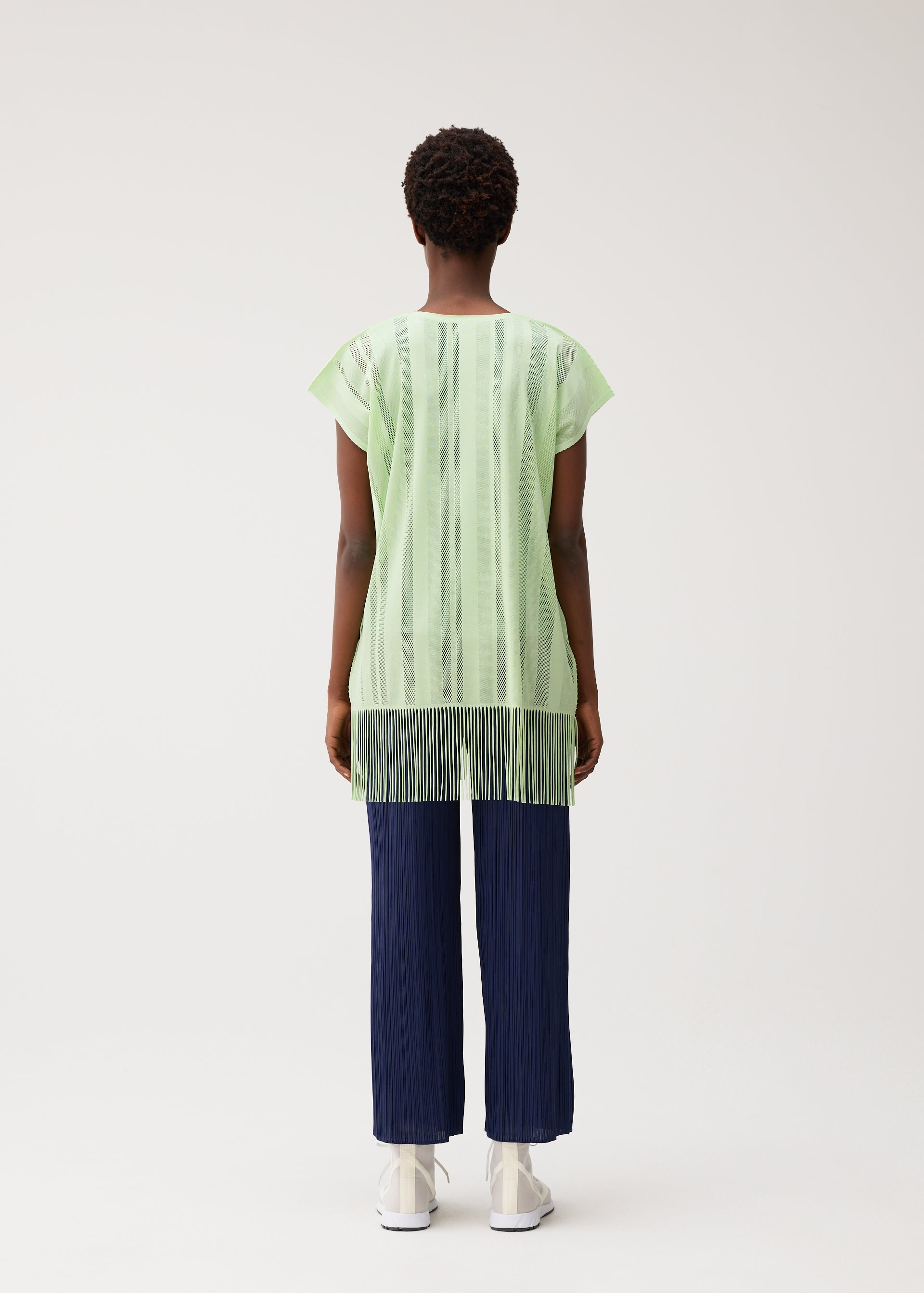 Zoomed view A person stands facing away against a plain light background, wearing the PLEATS PLEASE ISSEY MIYAKE A-POC LACE short-sleeve fringed tunic with vertical stripes in light green, paired with navy blue pleated pants and white shoes.