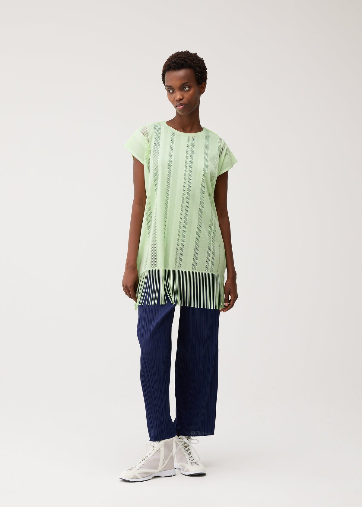 A person wearing the PLEATS PLEASE ISSEY MIYAKE A-POC LACE light green, round-neck tunic featuring vertical stripes and fringe at the hem, paired with navy blue pleated pants and white lace-up shoes, standing against a plain light background. view 8 of 9