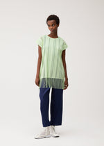 A person wearing the PLEATS PLEASE ISSEY MIYAKE A-POC LACE light green, round-neck tunic featuring vertical stripes and fringe at the hem, paired with navy blue pleated pants and white lace-up shoes, standing against a plain light background.