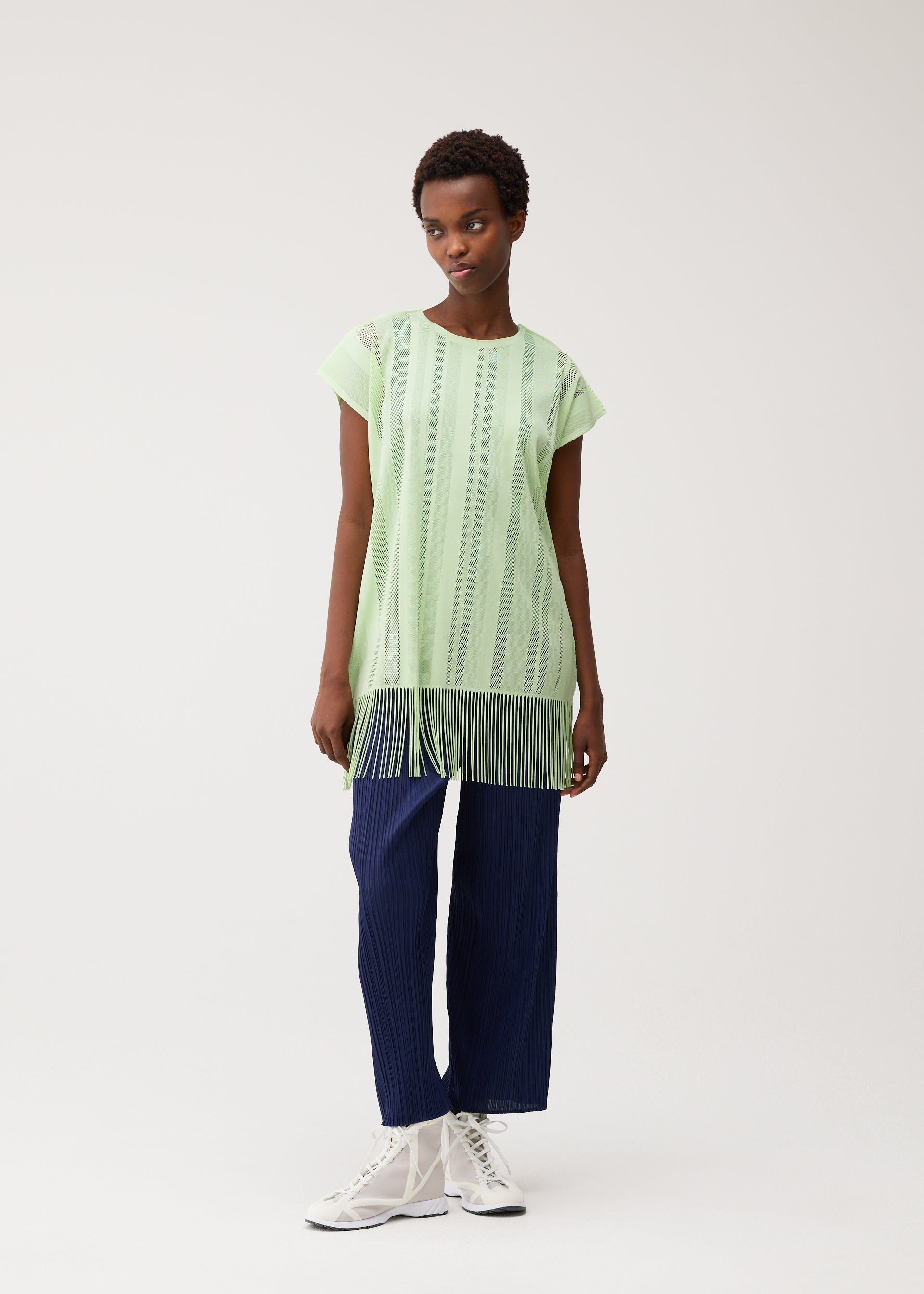 Zoomed view A person wearing the PLEATS PLEASE ISSEY MIYAKE A-POC LACE light green, round-neck tunic featuring vertical stripes and fringe at the hem, paired with navy blue pleated pants and white lace-up shoes, standing against a plain light background.