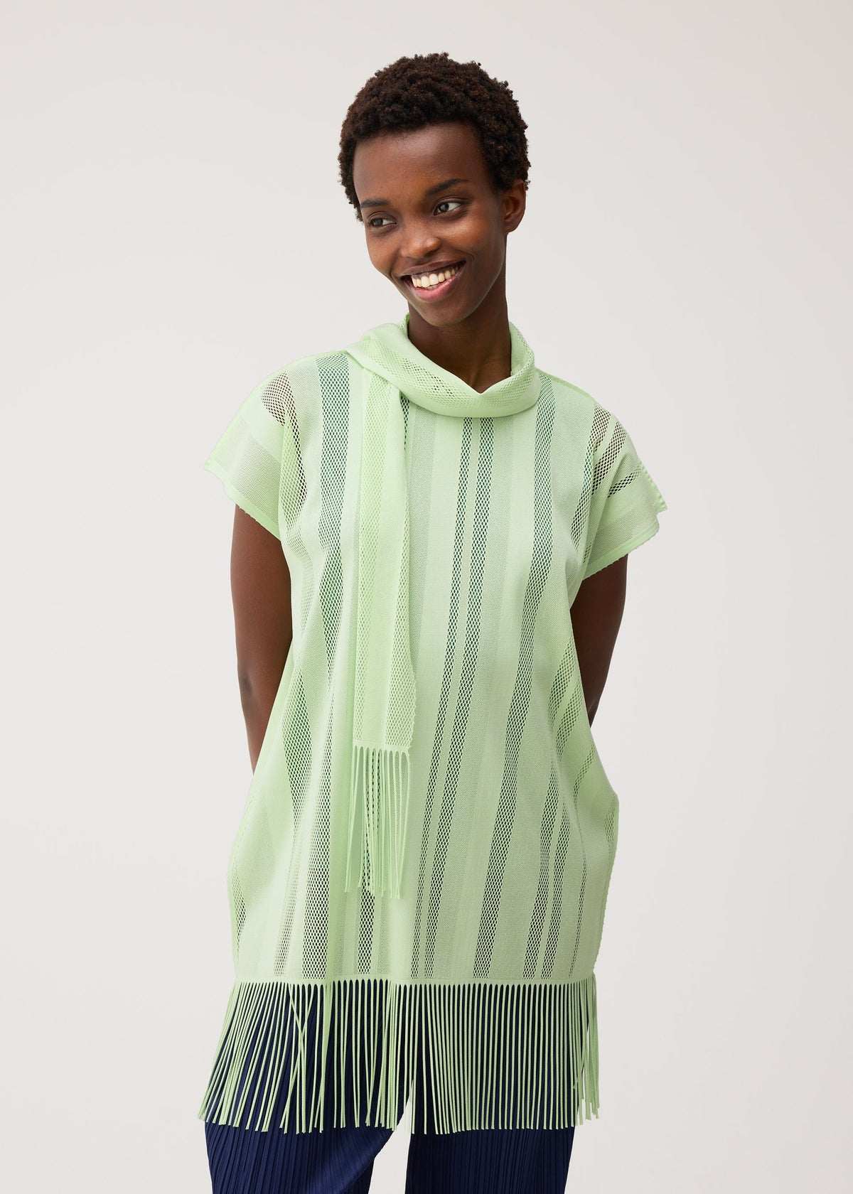 A person wearing the PLEATS PLEASE ISSEY MIYAKE A-POC LACE—a light green, short-sleeved tunic with vertical stripes, a cowl neck, and fringed edges—smiles with hands behind their back against a light background. The top’s delicate mesh material lends an airy touch to the overall look. view 9 of 9