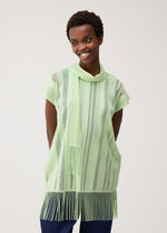 A person wearing the PLEATS PLEASE ISSEY MIYAKE A-POC LACE—a light green, short-sleeved tunic with vertical stripes, a cowl neck, and fringed edges—smiles with hands behind their back against a light background. The top’s delicate mesh material lends an airy touch to the overall look.