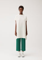 A person with short hair and a neutral expression stands against a plain background wearing the PLEATS PLEASE ISSEY MIYAKE PAPER BOUNCE KNIT—a loose, white washi-paper-blend high-neck tunic—over green pleated pants and white high-top sneakers.