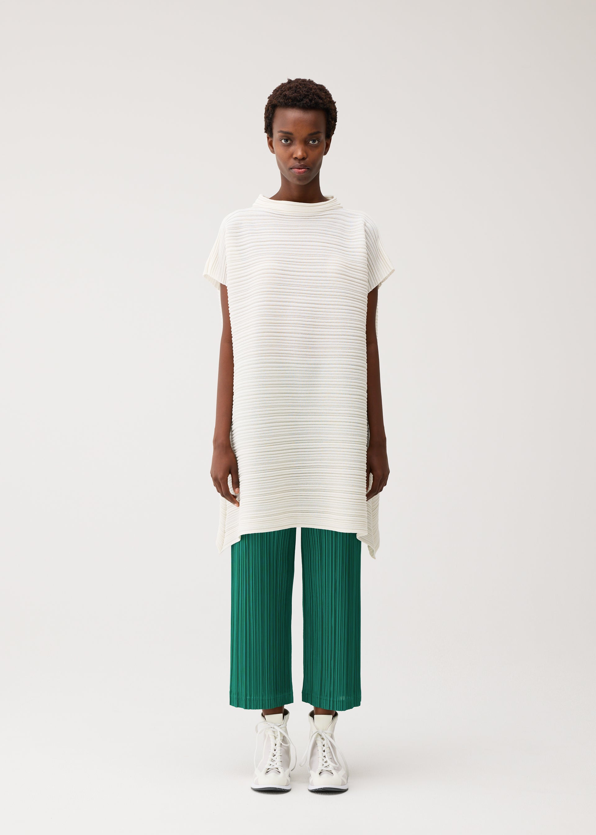 Zoomed view A person with short hair and a neutral expression stands against a plain background wearing the PLEATS PLEASE ISSEY MIYAKE PAPER BOUNCE KNIT—a loose, white washi-paper-blend high-neck tunic—over green pleated pants and white high-top sneakers.