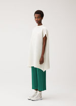 A woman stands against a plain background wearing the PAPER BOUNCE KNIT tunic in washi-paper-blend and bright green pleated pants from PLEATS PLEASE ISSEY MIYAKE, paired with white high-top sneakers.