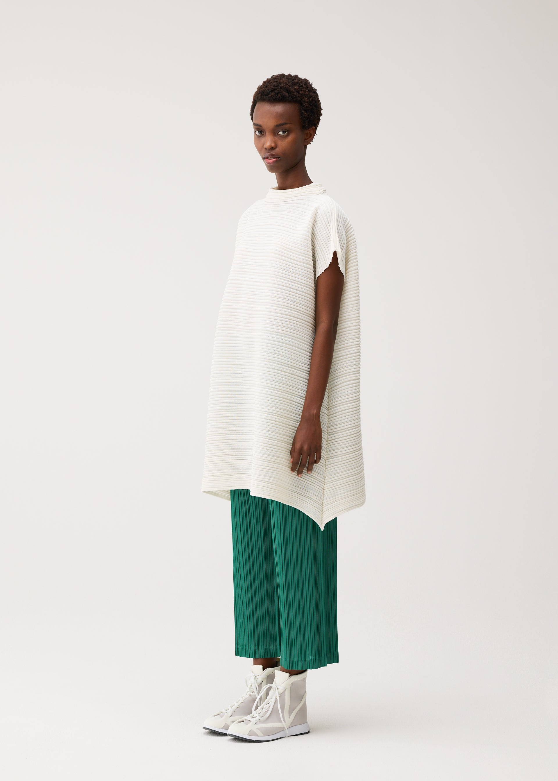 Zoomed view A woman stands against a plain background wearing the PAPER BOUNCE KNIT tunic in washi-paper-blend and bright green pleated pants from PLEATS PLEASE ISSEY MIYAKE, paired with white high-top sneakers.