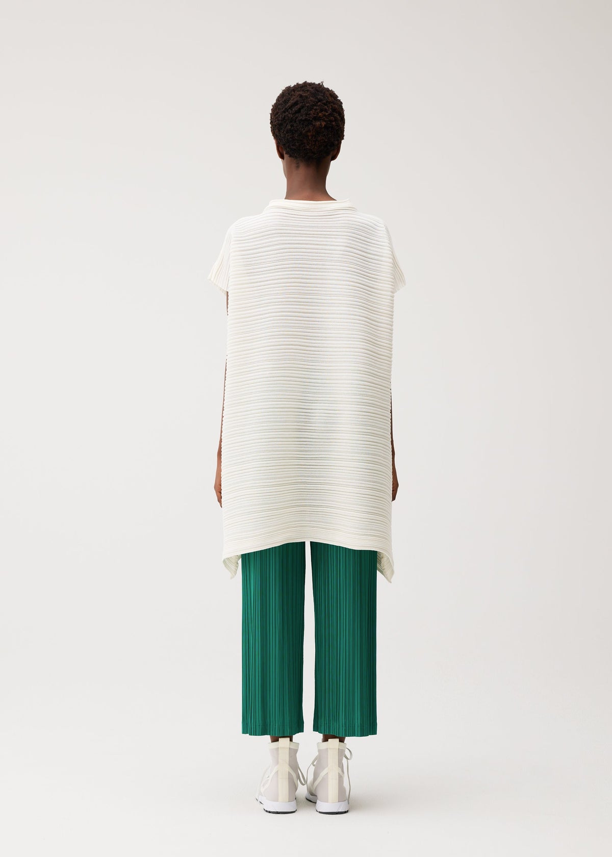 A person faces away, wearing PLEATS PLEASE ISSEY MIYAKE's PAPER BOUNCE KNIT—a loose, short-sleeve white washi-paper-blend top with subtle stripes—green pleated knit cropped pants, and white shoes against a light background. view 7 of 8