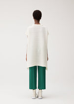 A person faces away, wearing PLEATS PLEASE ISSEY MIYAKE's PAPER BOUNCE KNIT—a loose, short-sleeve white washi-paper-blend top with subtle stripes—green pleated knit cropped pants, and white shoes against a light background.