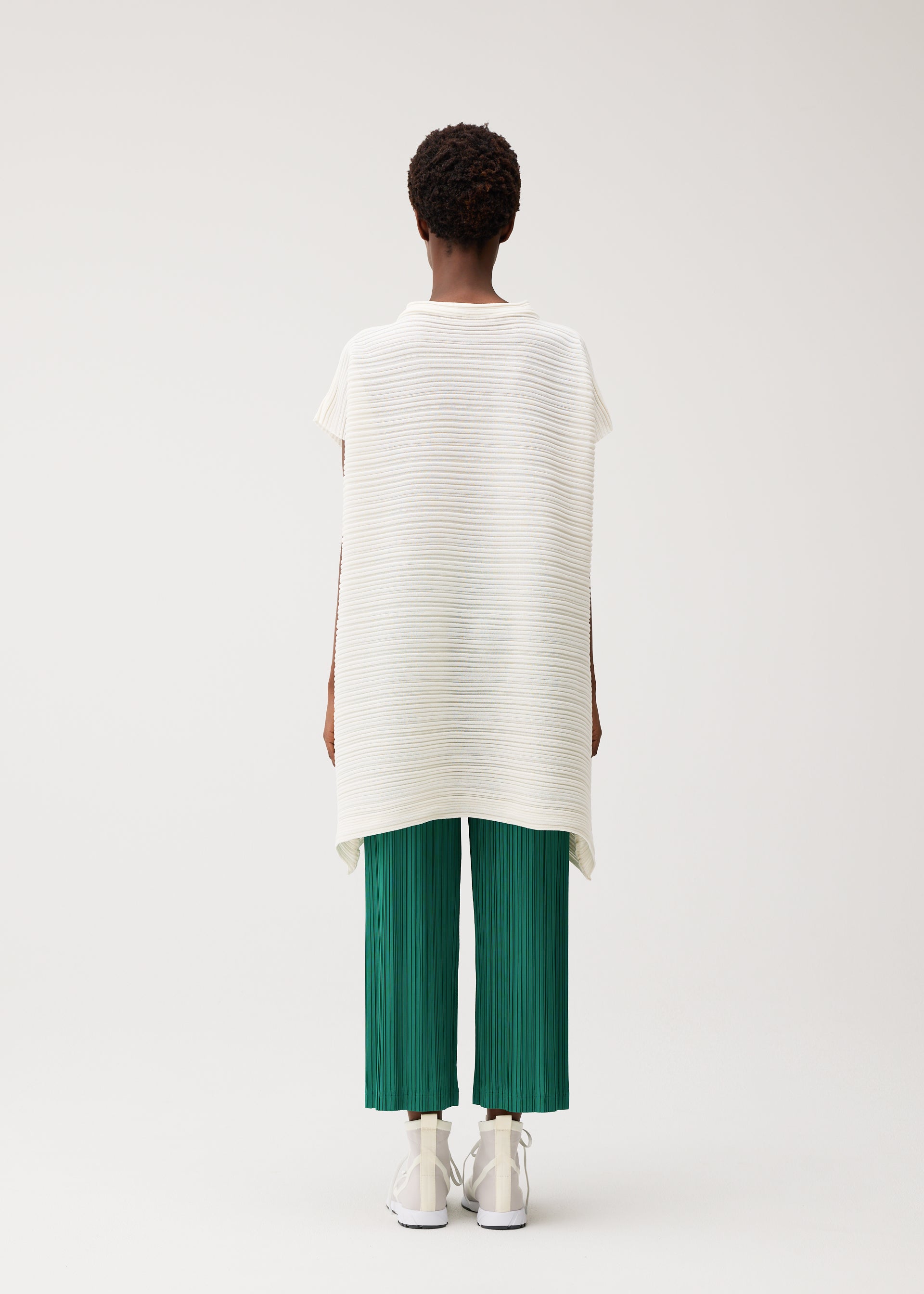 Zoomed view A person faces away, wearing PLEATS PLEASE ISSEY MIYAKE's PAPER BOUNCE KNIT—a loose, short-sleeve white washi-paper-blend top with subtle stripes—green pleated knit cropped pants, and white shoes against a light background.