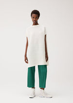 A person stands against a plain background wearing PLEATS PLEASE ISSEY MIYAKE’s PAPER BOUNCE KNIT: a loose, off-white, washi-paper-blend high-neck tunic with horizontal texture and green pleated knit wide-leg pants, paired with white high-top sneakers.