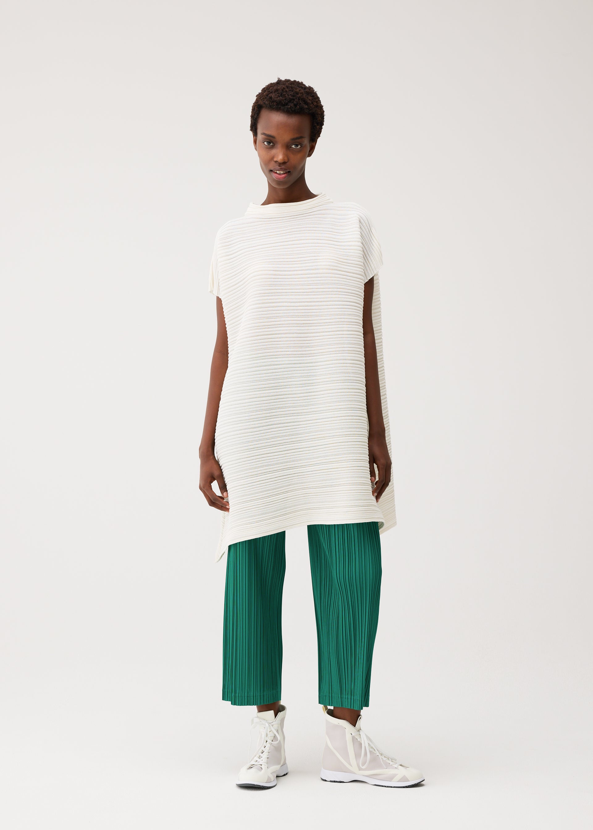 Zoomed view A person stands against a plain background wearing PLEATS PLEASE ISSEY MIYAKE’s PAPER BOUNCE KNIT: a loose, off-white, washi-paper-blend high-neck tunic with horizontal texture and green pleated knit wide-leg pants, paired with white high-top sneakers.