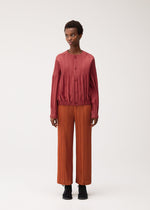 A person stands facing forward against a plain white background, dressed for chilly seasons in a textured, long-sleeve red top and loose orange pleated pants made from thick fabric, completed with black shoes. The look features the SMOOTH COAT by PLEATS PLEASE ISSEY MIYAKE.