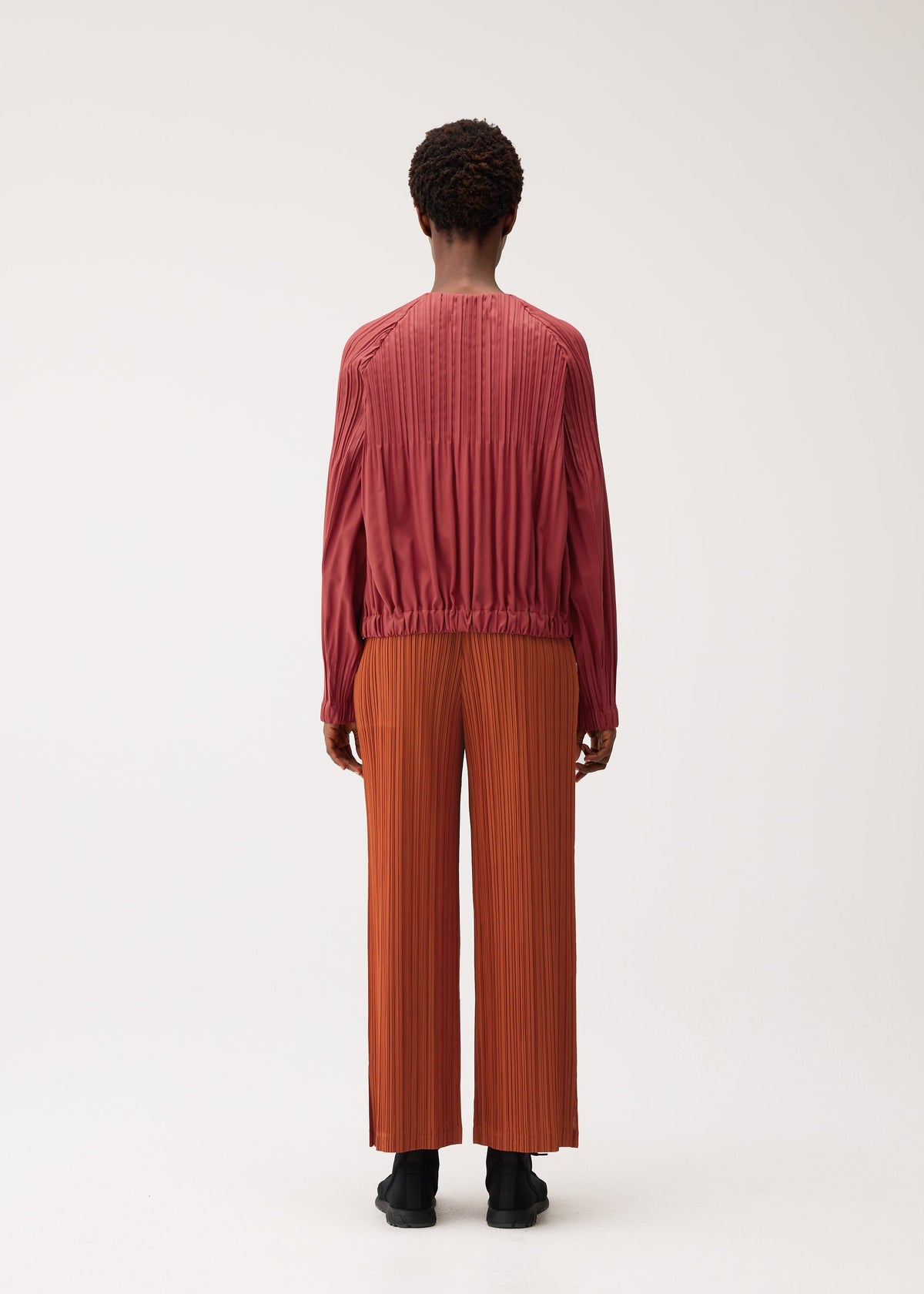 A person stands with their back to the camera against a plain light background, wearing the SMOOTH COAT by PLEATS PLEASE ISSEY MIYAKE—a long-sleeved, rose-colored pleated top paired with wide-leg, rust-orange pleated pants crafted from thick fabric that’s perfect for chilly seasons. view 7 of 8