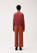 A person stands with their back to the camera against a plain light background, wearing the SMOOTH COAT by PLEATS PLEASE ISSEY MIYAKE—a long-sleeved, rose-colored pleated top paired with wide-leg, rust-orange pleated pants crafted from thick fabric that’s perfect for chilly seasons.