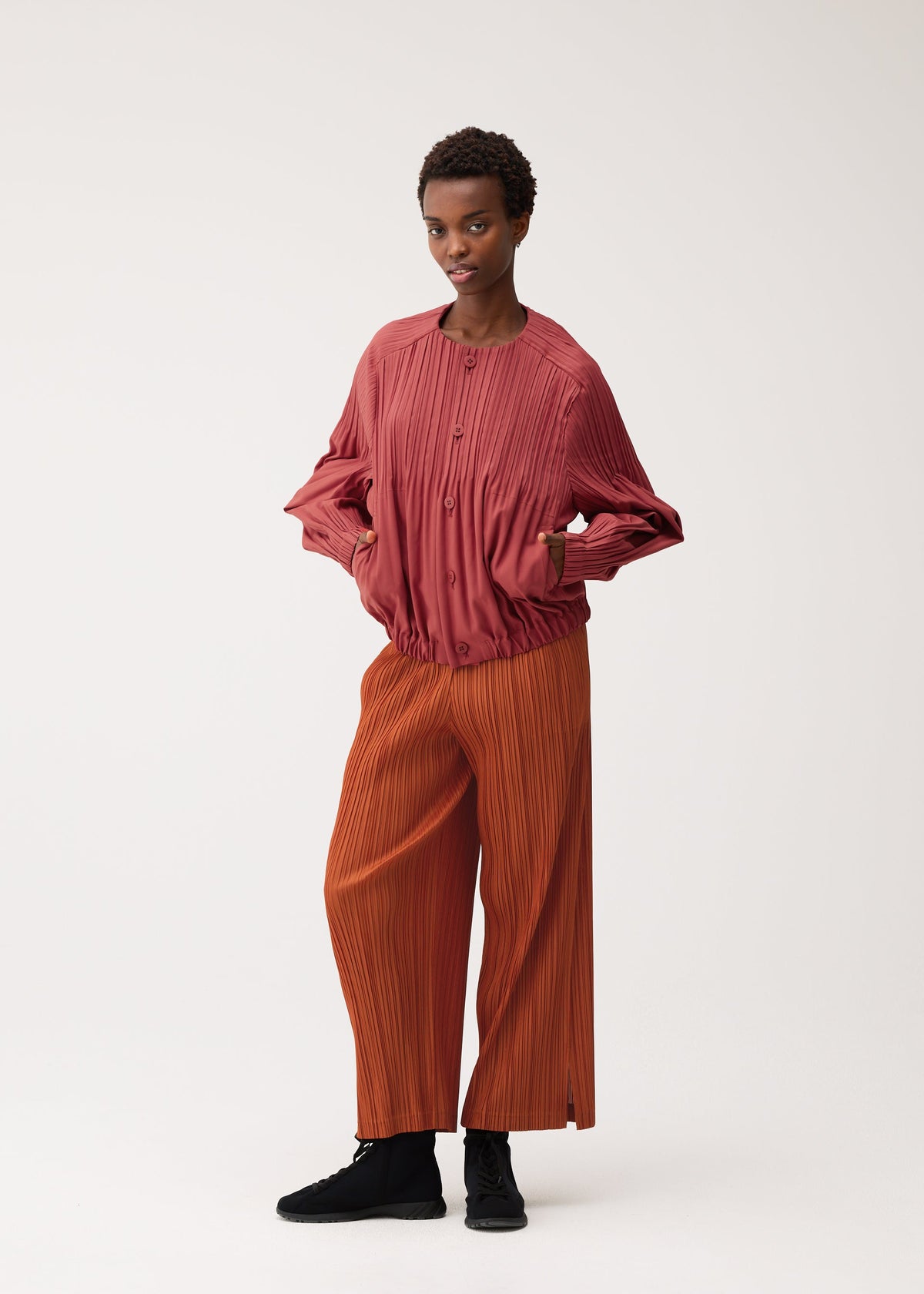 A person stands against a plain backdrop, hands in pockets and wearing a SMOOTH COAT by PLEATS PLEASE ISSEY MIYAKE—a textured red button-up outerwear piece with long sleeves—paired with loose, thick orange pleated pants and black shoes. With a neutral expression, the outfit is ideal for cooler weather. view 8 of 8