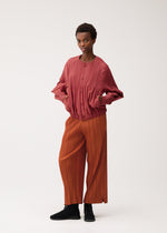 A person stands against a plain backdrop, hands in pockets and wearing a SMOOTH COAT by PLEATS PLEASE ISSEY MIYAKE—a textured red button-up outerwear piece with long sleeves—paired with loose, thick orange pleated pants and black shoes. With a neutral expression, the outfit is ideal for cooler weather.