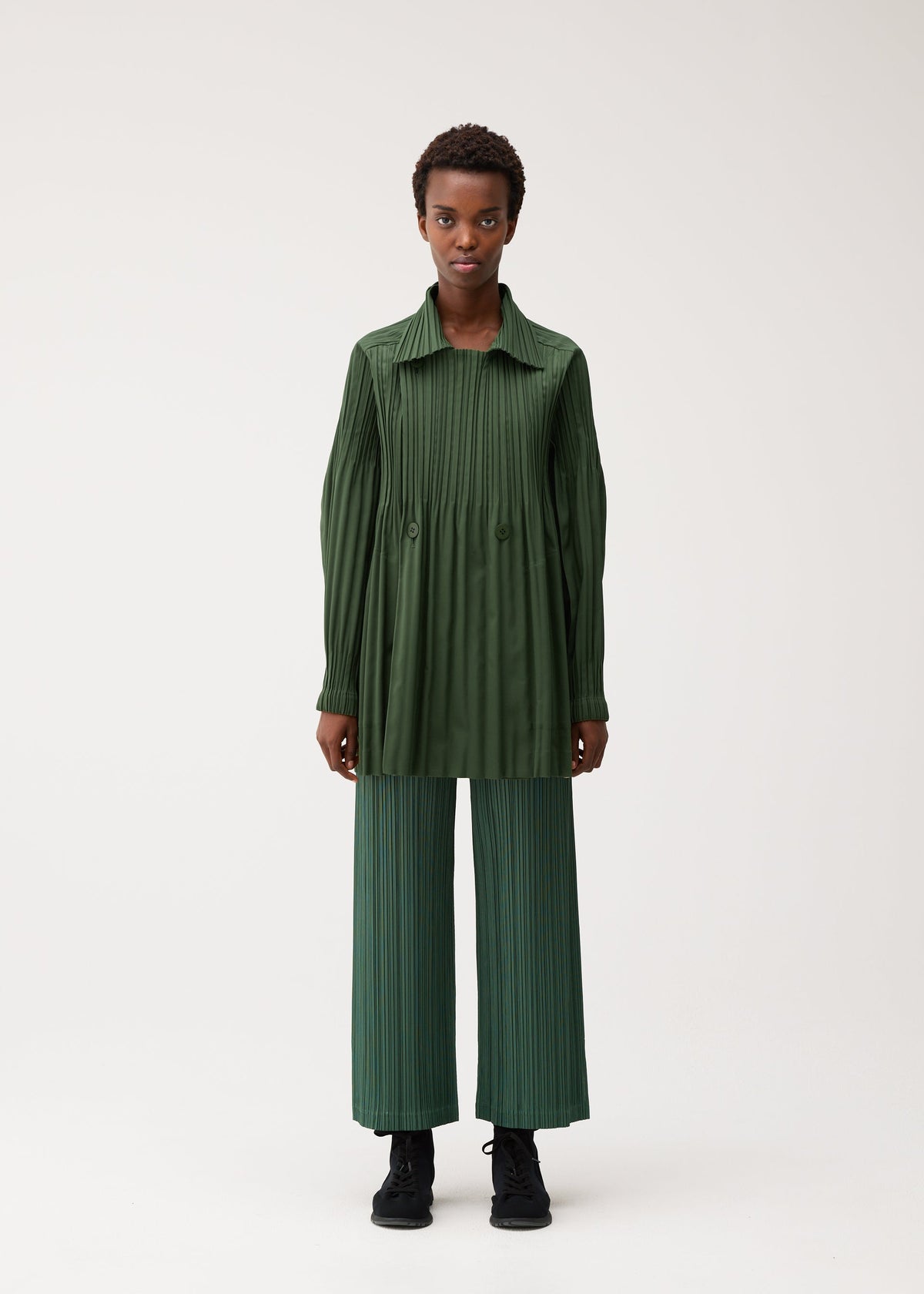 A person faces forward wearing the SMOOTH COAT from PLEATS PLEASE ISSEY MIYAKE—a long, pleated, dark green top made from thick fabric—paired with wide-legged, pleated, light green pants and black shoes against a plain light background, creating a stylish and comfortable look ideal for chilly seasons. view 5 of 9