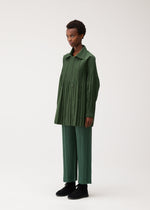 A person stands against a plain background, wearing a long, pleated green top and matching pleated green pants with black shoes. Layered over their outfit is the SMOOTH COAT by PLEATS PLEASE ISSEY MIYAKE—a collarless short coat crafted from thick fabric, making it an ideal outerwear choice for chilly seasons. They face slightly to the left with a neutral expression.