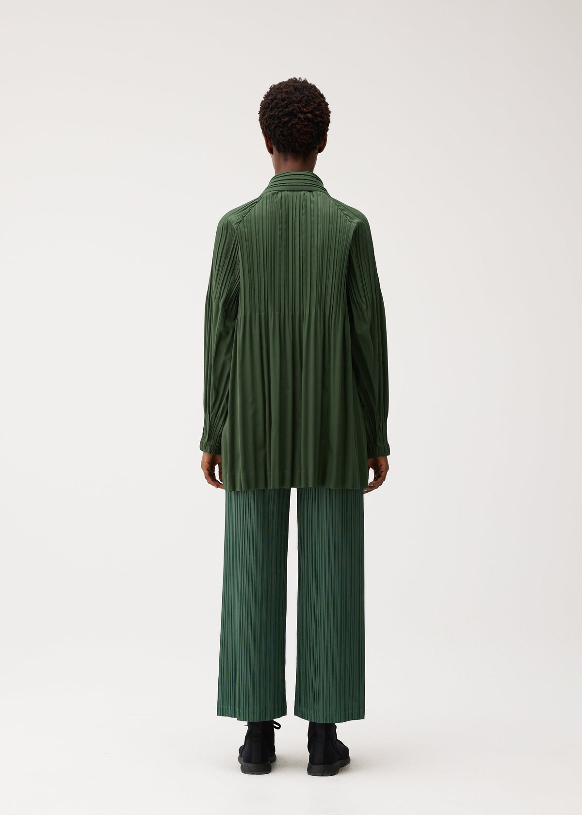 A person stands facing away against a plain white background, wearing PLEATS PLEASE ISSEY MIYAKE’s SMOOTH COAT—a collarless short coat crafted from thick fabric—layered over a loose, long-sleeved dark green shirt and wide-legged pleated green pants with black shoes, creating an ideal look for the chilly season. view 7 of 9