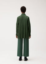 A person stands facing away against a plain white background, wearing PLEATS PLEASE ISSEY MIYAKE’s SMOOTH COAT—a collarless short coat crafted from thick fabric—layered over a loose, long-sleeved dark green shirt and wide-legged pleated green pants with black shoes, creating an ideal look for the chilly season.