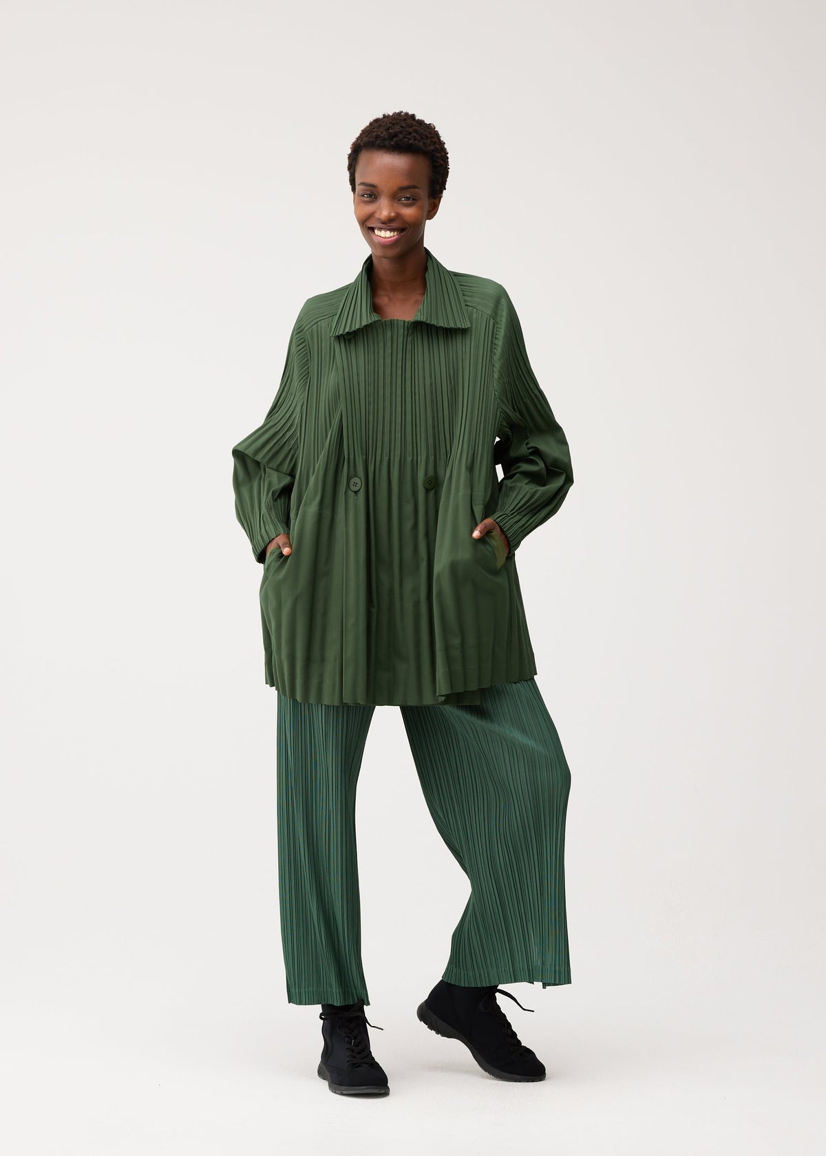 Zoomed view A person smiles while wearing the SMOOTH COAT by PLEATS PLEASE ISSEY MIYAKE—a green pleated jacket crafted from thick fabric—paired with matching wide-leg pleated pants and black shoes, standing against a plain light background. This ensemble is perfect attire for chilly seasons.