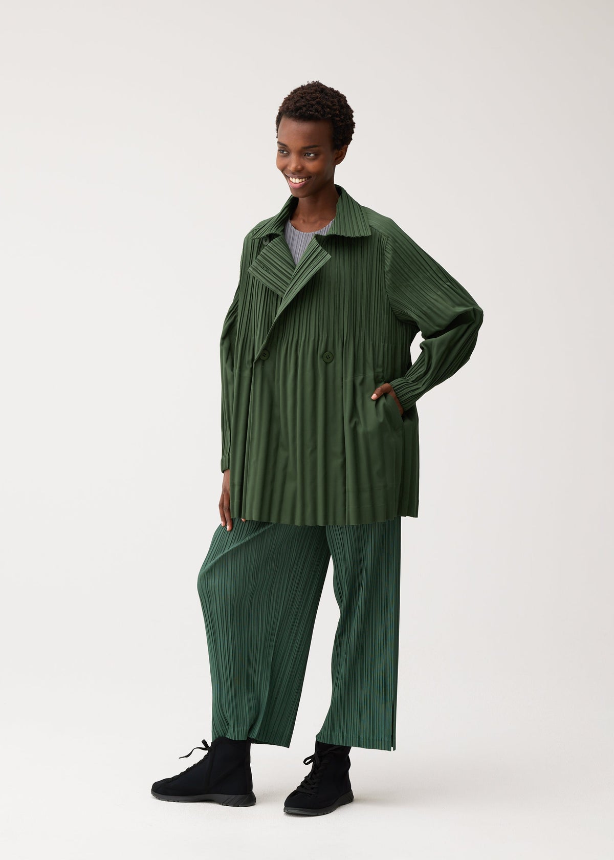 Zoomed view A person stands smiling in front of a plain, light background, dressed in the PLEATS PLEASE ISSEY MIYAKE SMOOTH COAT—a matching loose green pleated jacket and pants set crafted from thick fabric ideal for chilly seasons. The collarless short coat lends a modern edge, paired with black shoes and a gray top.