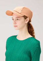 A young woman with fair skin and red hair tied back wears the PLEATS PLEASE ISSEY MIYAKE PLEATS CAP and a green pleated top, gazing to the side against a plain light background.