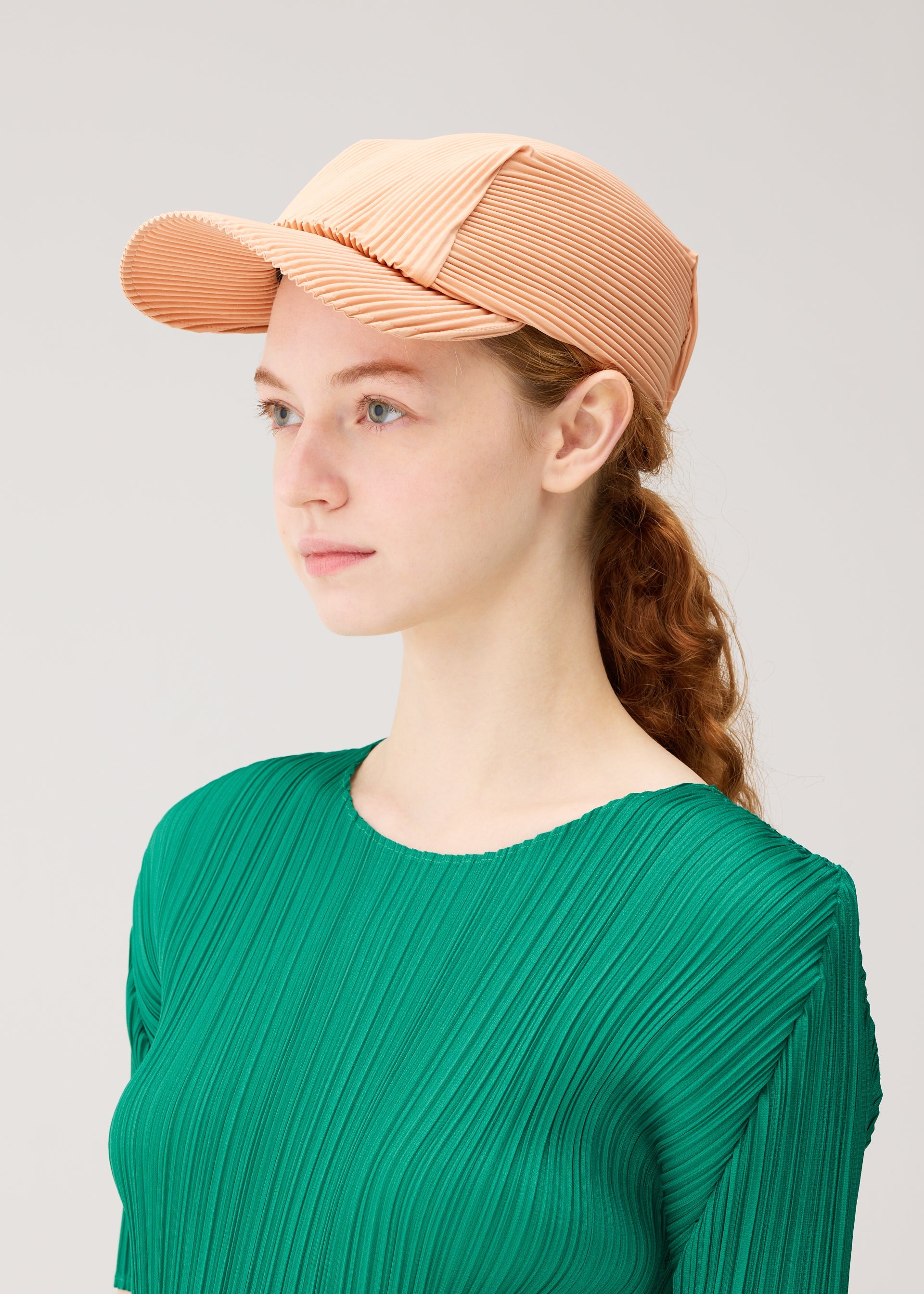 Zoomed view A young woman with fair skin and red hair tied back wears the PLEATS PLEASE ISSEY MIYAKE PLEATS CAP and a green pleated top, gazing to the side against a plain light background.