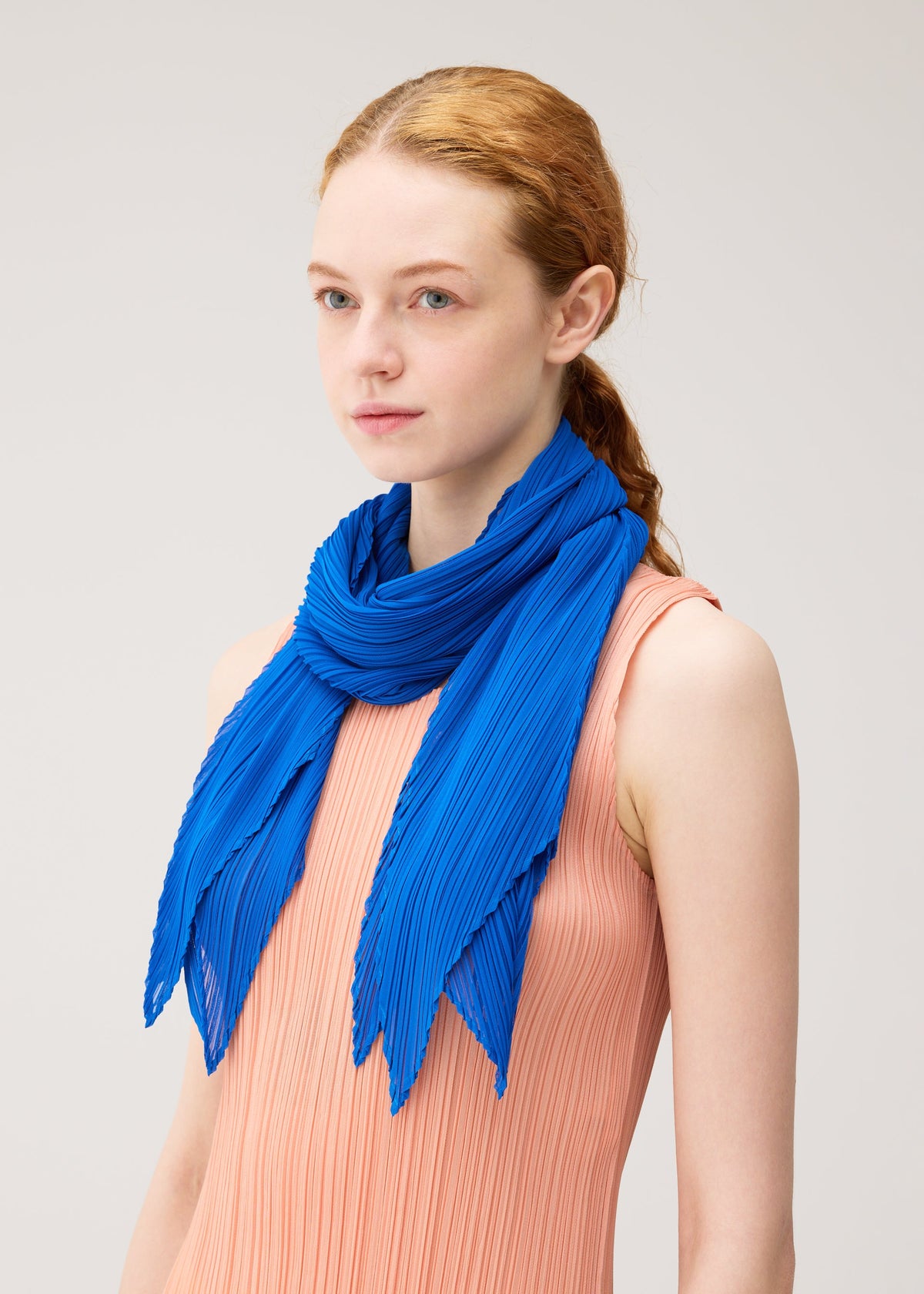 A young woman with fair skin and red hair wears a sleeveless peach pleated dress and the MONTHLY SCARF JULY by PLEATS PLEASE ISSEY MIYAKE, styled in vibrant pop-art colors against a plain light background. view 6 of 6