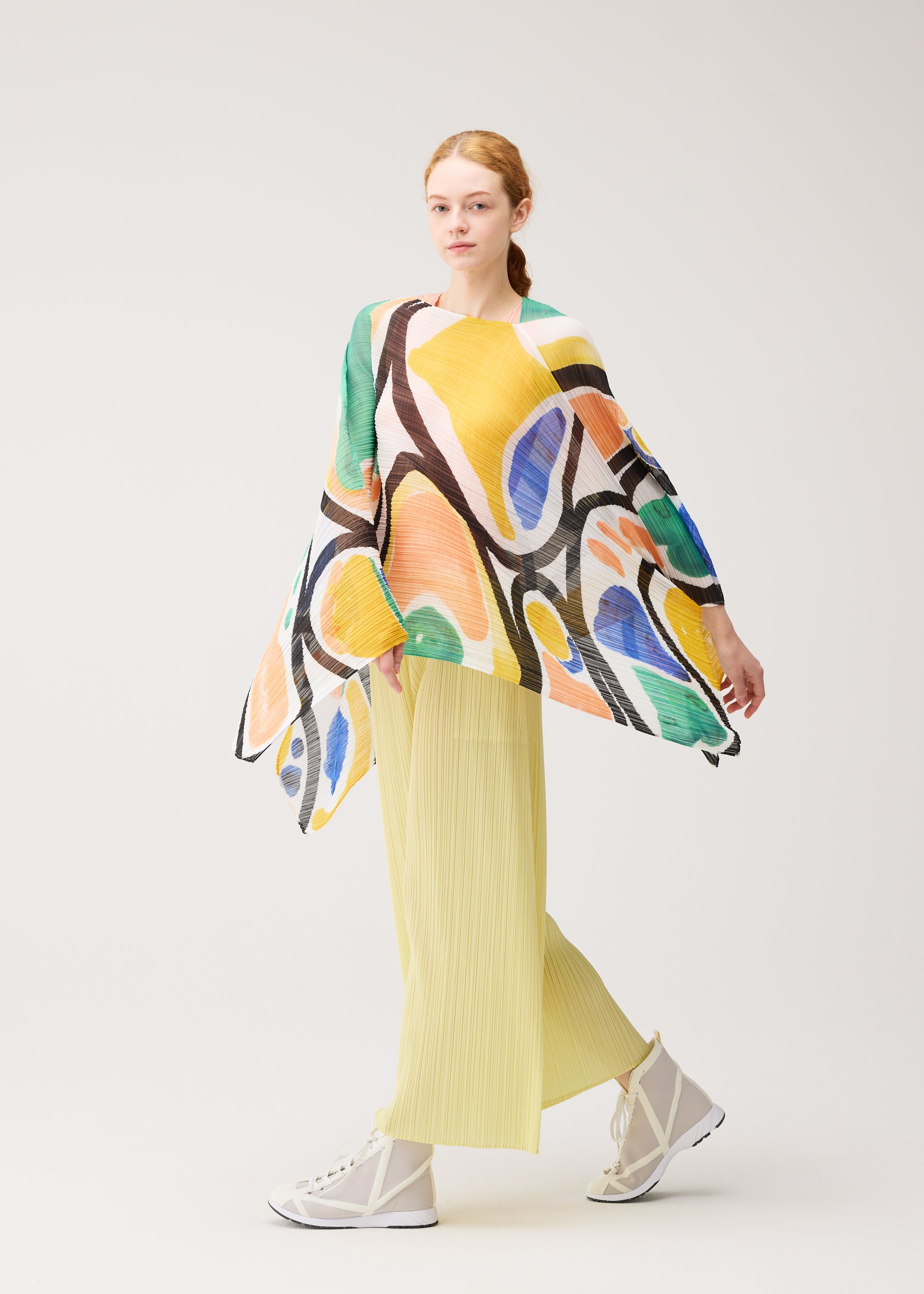 Zoomed view A person with light skin and red hair models the ROLL MADAME-T by PLEATS PLEASE ISSEY MIYAKE: a vibrant abstract-patterned top with wide sleeves, a pleated stole, light yellow pleated pants, and white high-top shoes against a plain background.