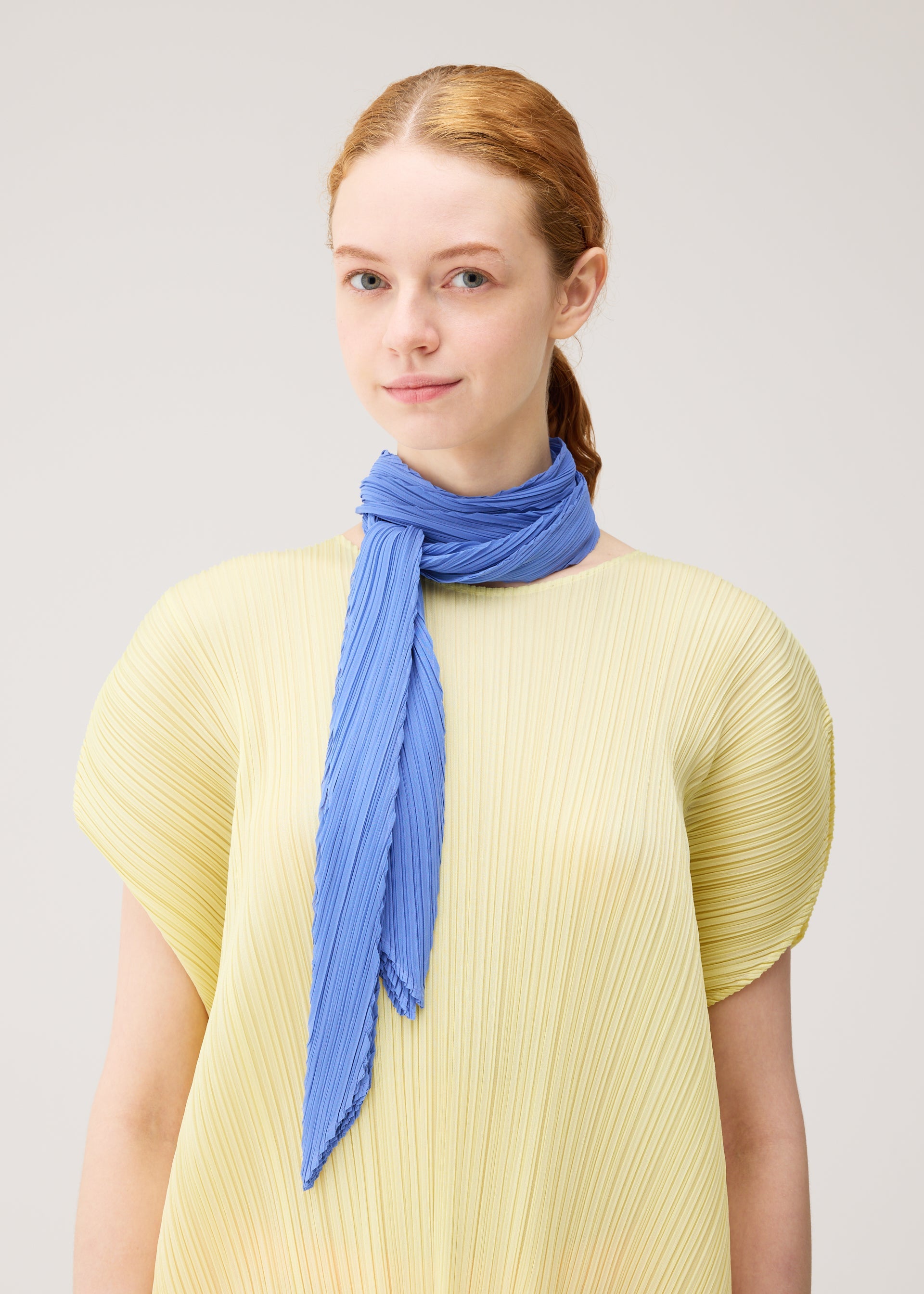 Zoomed view A young woman with fair skin and red hair wears a pleated light yellow top and the PLEATS PLEASE ISSEY MIYAKE SKINNY SCARF in blue, wrapped around her neck, standing against a plain light background.