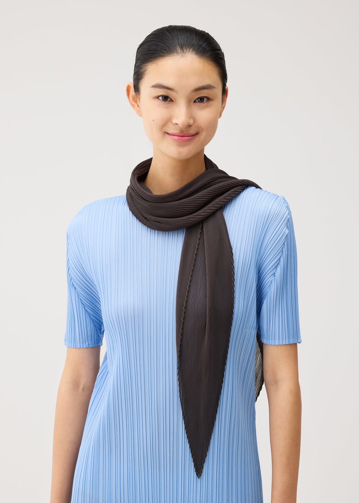 A person is wearing the PLEATS PLEASE ISSEY MIYAKE MONTHLY SCARF AUGUST—a diamond-shaped dark brown scarf draped stylishly around their neck—paired with a light blue pleated short-sleeve dress. They are smiling at the camera against a plain light background, and the refreshing colors of the outfit enhance its lively midsummer vibe. view 5 of 5