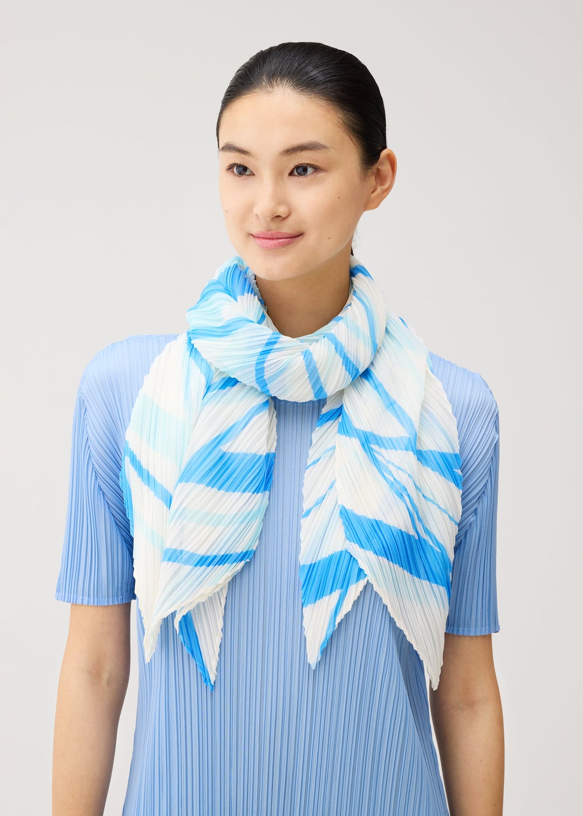 A woman with dark hair pulled back wears a light blue, pleated, short-sleeved top and drapes the PLEATS PLEASE ISSEY MIYAKE FOLD LINE SCARF—a matching blue and white striped, pleated scarf—loosely around her neck, its delicate folds reminiscent of a folding screen, set against a plain light gray background. view 5 of 5