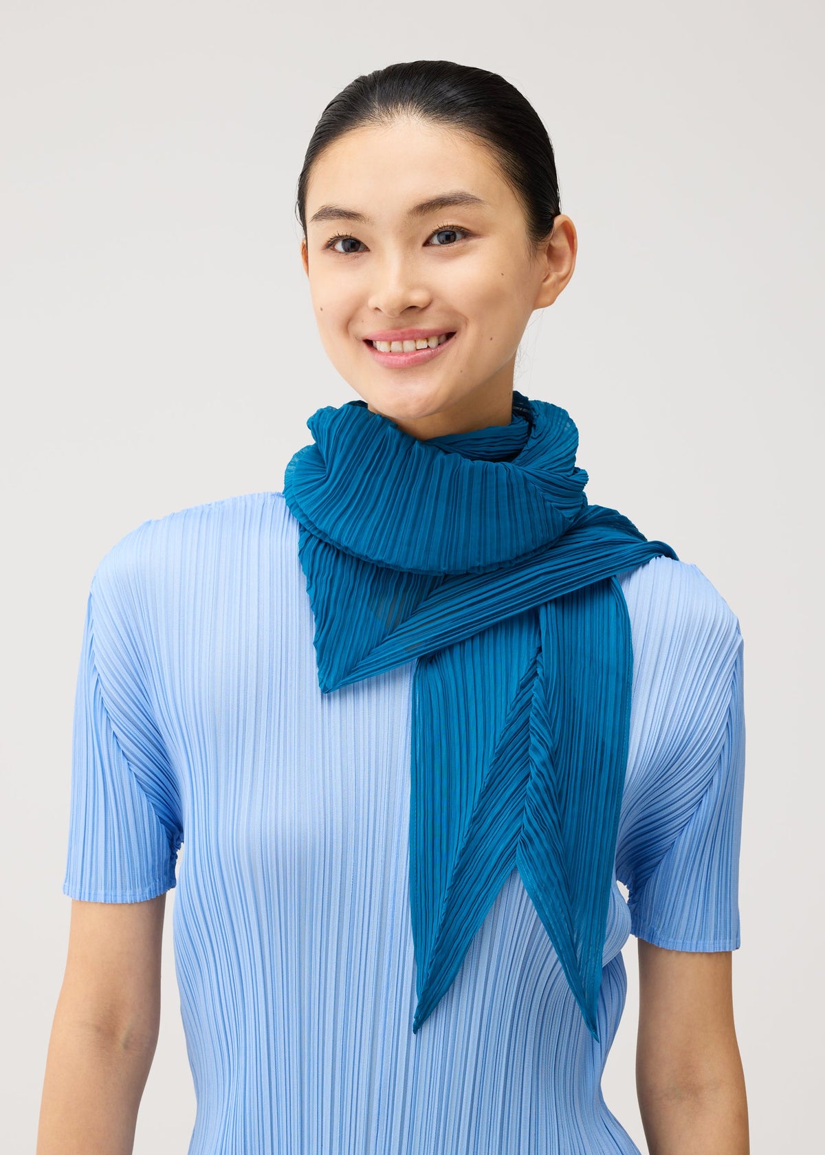 A person with dark hair tied back, wearing a light blue pleated top and the FOLD HOOD SCARF by PLEATS PLEASE ISSEY MIYAKE in teal, smiles against a plain light background. view 5 of 6