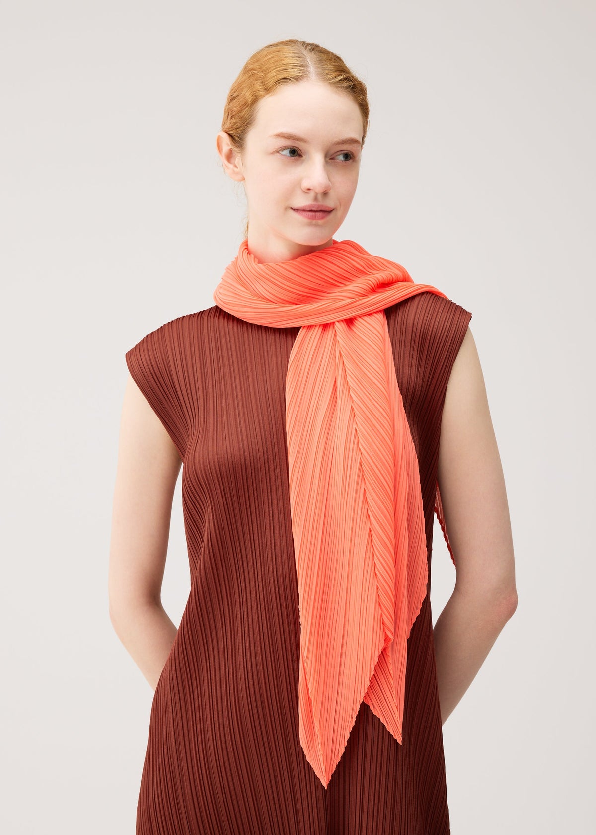 A person with light skin and red hair, wearing a brown pleated sleeveless dress and the PLEATS PLEASE ISSEY MIYAKE MONTHLY SCARF SEPTEMBER—a bright coral folding screen scarf—wrapped around their neck, stands against a plain light background. The gentle colors of the outfit create an elegant, understated effect. view 6 of 6
