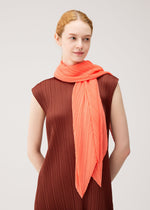 A person with light skin and red hair, wearing a brown pleated sleeveless dress and the PLEATS PLEASE ISSEY MIYAKE MONTHLY SCARF SEPTEMBER—a bright coral folding screen scarf—wrapped around their neck, stands against a plain light background. The gentle colors of the outfit create an elegant, understated effect.