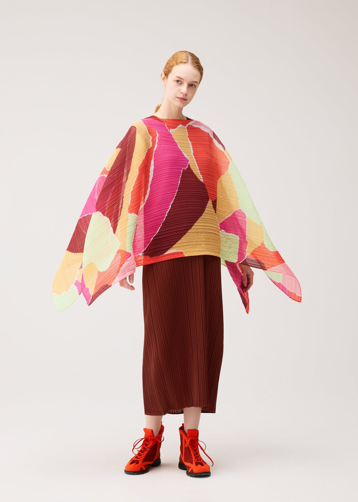 A woman poses against a plain light background wearing the SURPRISE! MADAME-T top by PLEATS PLEASE ISSEY MIYAKE, which features a colorful, geometric-inspired design with a subtle paper tearing pattern and wide sleeves, paired with a pleated brown skirt and bright orange boots. view 4 of 4