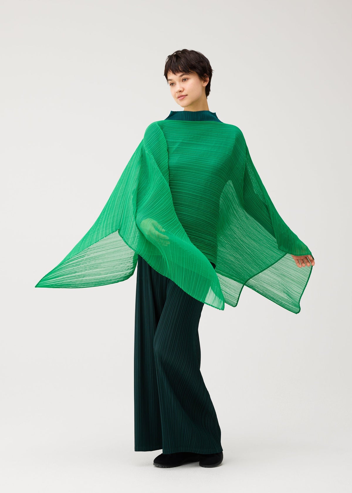 Zoomed view Wearing the MADAME-T OCTOBER by PLEATS PLEASE ISSEY MIYAKE, a person in a bright green pleated stole top and dark green wide-leg pants stands against a plain background, looking to the side.